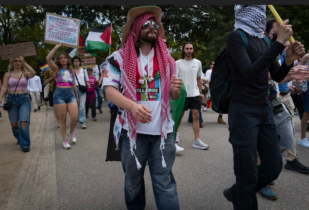 Foto van een bebaarde man in BIJ1 t-shirt met ene rode lkeffiye om. P de achtergrond demonstratnen voor Palestina en trans rechten op de Pride March